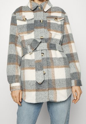 Wełniana kurtka na długi rękaw z paskiem, w kratę w odcieniach szarości, bieli i beżu, zapinana na guziki z przodu, z kieszeniami na piersi, noszona na niebieskich dżinsach.