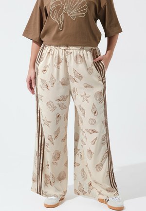 Pantalon ample crème avec imprimés de coquillages marron et d'étoiles de mer, taille élastique, et bandes latérales marron, porté avec un haut marron et des baskets blanches.