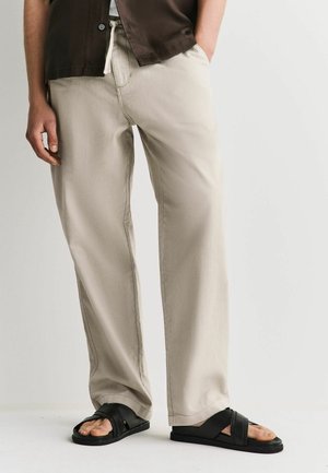 Person trägt beige lockere Hose und schwarzes Hemd, steht mit einer Hand in der Tasche, schwarze offene Sandalen auf weißem Boden.