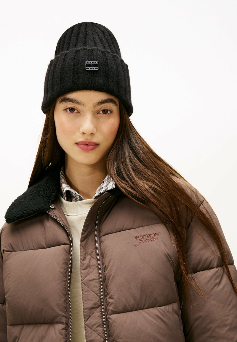 Zwarte geribbelde gebreide beanie met logo-opdruk, gedragen met een bruine pufferjack die een zwarte schapenvachtkraag heeft en een crème trui eronder.