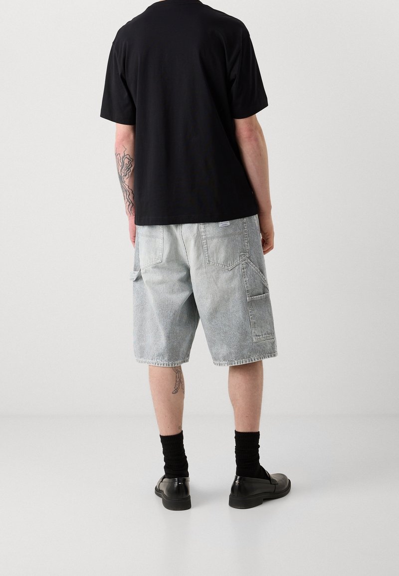 Homme portant une chemise noire à manches courtes, un short gris clair au genou, des chaussettes noires et des chaussures noires, debout dos à la caméra.