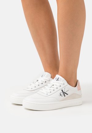 Baskets basses blanches avec logo "CK Calvin Klein Jeans" sur le côté, portées sur pieds nus, avec un accent rose au talon et des lacets blancs.