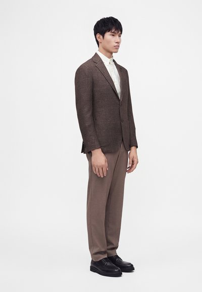 Jeune homme portant un blazer marron à carreaux, une chemise de couleur claire, un pantalon marron et des chaussures habillées noires, debout devant un fond blanc uni.