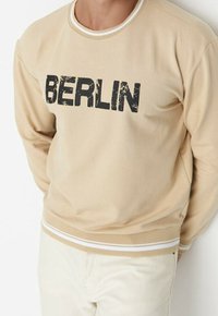 Sudadera beige con el texto desgastado en negro "BERLÍN", llevada por una persona con pantalones claros, recortada de hombros a caderas.