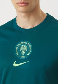 Teal athletic shirt with a round neck, featuring a Nigeria Football Federation logo and a green Nike swoosh on the chest.