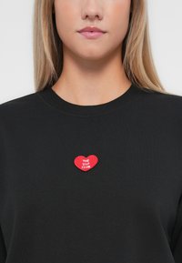 Hanorac negru cu un patch în formă de inimă roșie, pe care este inscripționat cu alb "THE V.I.P. CLUB" în zona pieptului. Textură moale, guler rotund.