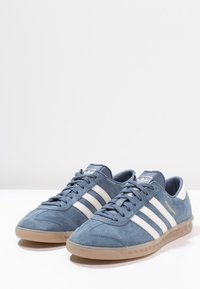 Blue suede athletic shoes with white stripes and branding, featuring a gum sole and textured upper. Lace-up design with reinforced toe.