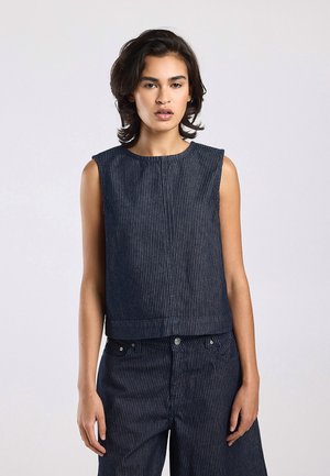 Femme portant un top sans manches bleu marine à fines rayures et un pantalon large assorti, debout devant un fond clair uni.