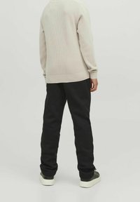 Man i beige tröja, svarta byxor och svarta sneakers med vita sulor, står med ryggen mot kameran mot en enfärgad ljus bakgrund.