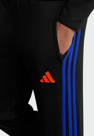 Mano en el bolsillo de pantalones negros con rayas laterales azules y logo triangular rojo cerca de la cremallera del bolsillo.