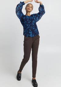 Blusa floreale blu navy con maniche a palloncino trasparenti, abbinata a pantaloni aderenti marroni e mocassini neri, che mette in risalto uno stile a strati.