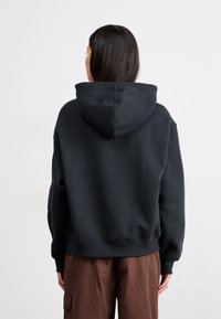 Sweat à capuche noir, surdimensionné, avec une texture douce, des poignets et un ourlet côtelés. Porté sur un pantalon cargo marron, vu de dos.