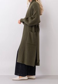 Oliwkowy, prążkowany kardigan o luźnym kroju, z szerokimi rękawami i bocznymi kieszeniami; związany pasek w talii, zestawiony z ciemnymi spodniami o szerokich nogawkach i białymi butami.