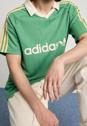 Osoba ubrana w zieloną koszulkę Adidas z białym kołnierzykiem i żółtymi paskami na rękawach, w połączeniu z jasnobeżowymi spodniami, siedząca na niebieskiej podłodze.