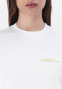 Bílý bavlněný tričko s kulatým výstřihem a krátkými rukávy, ozdobené zlatě vyšitým logem "BARBOUR INTERNATIONAL" na hrudníku.