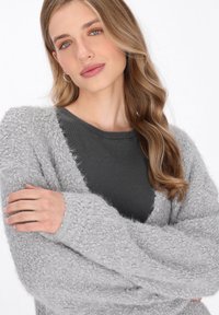 Grauer, fluffiger Cardigan mit lockeren Ärmeln und einer strukturierten Oberfläche, getragen über einem dunkelgrauen Rippenshirt. Glattes Haar fällt über eine Schulter.