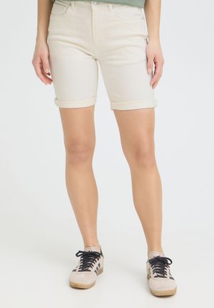 Persona que lleva puestos unos pantalones cortos vaqueros beige claro con dobladillo enrollado y zapatillas beige con cordones negros, de pie sobre un fondo blanco liso.