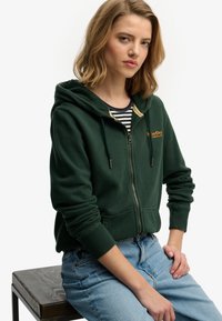 Sudadera verde con cremallera hecha de tela suave con capucha, con un logo dorado en el pecho izquierdo, combinada con vaqueros azul claro.