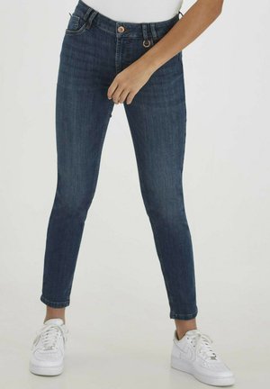 Jeans ajustados de denim en azul oscuro con un ligero acabado desteñido, diseño de cinco bolsillos y cierre de botón en la cintura. Combinados con zapatillas blancas.