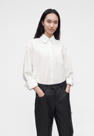 Femme aux longs cheveux foncés, portant une chemise blanche à boutons avec manches retroussées et un pantalon en cuir noir, debout les mains dans les poches.