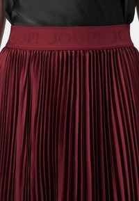 Burgundy pleated skirt with a wide waistband featuring the embossed logo "JOOP!" in a repeating pattern. Smooth texture and flowing design.