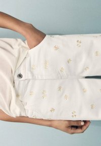 Pantalons blancs avec broderies florales jaunes, présentant une coupe décontractée et des poches latérales. Le matériau semble texturé avec un léger éclat.