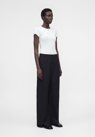 Γυναίκα όρθια, φοράει εφαρμοστή λευκή κοντομάνικη μπλούζα, φαρδιά μαύρη παντελόνα και μαύρα μοκασίνια μπροστά σε μονόχρωμο φόντο.