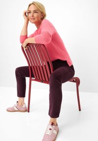 Magenta trui, donkerpaarse broek en lichtroze sneakers. Model zit op een rode, geribbelde metalen stoel tegen een witte achtergrond.