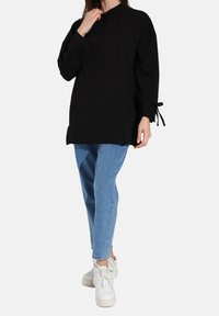 Pullover nera a maniche lunghe con collo alto e lacci ai polsini, abbinata a jeans dritti azzurri e sneakers bianche.