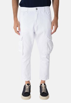 Uomo che indossa pantaloni cargo bianchi corti con tasche laterali e sneakers nere con lacci bianchi, in piedi davanti a uno sfondo semplice.