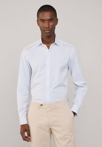 Camicia a righe blu chiaro con colletto classico, maniche lunghe e chiusura frontale con bottoni; abbinata a pantaloni beige.