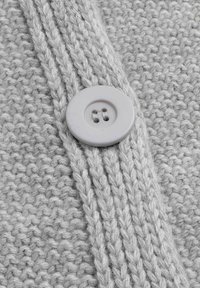 Gros plan d'un bouton blanc cousu sur un tissu tricoté gris clair texturé avec des motifs en côtes et en point de riz.