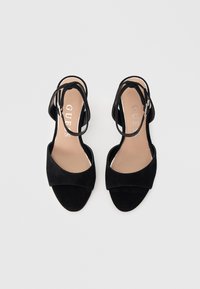 Zwarte suède sandalen met open teen, enkelriempjes en gespen, beige binnenzolen met GUESS-logo, getoond op een effen lichtgrijze achtergrond.