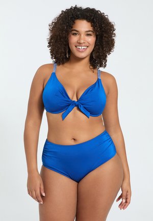 Krøllet-haired kvinde smilende, iført en blå bikini med bundet front top og høj talje underdel mod en ensfarvet hvid baggrund.