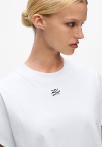 Tricou alb din bumbac, cu guler rotund și mâneci scurte. Prezintă un logo brodat negru în zona stânga a pieptului. Textură netedă.