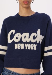 Marineblau geschnittenes Sweatshirt mit weißem "Coach New York"-Schriftzug, geripptem Rundhalsausschnitt und gestreiften Akzenten an den Ärmeln. Weicher, strukturierter Stoff.