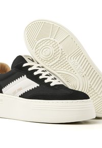 Black and white sneakers featuring a textured upper, flat laces, and a thick white sole with a patterned tread. Details include "EST. 1992" in gold.