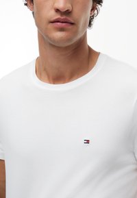 Young man wearing plain white t-shirt with small red, white, and blue logo on the left chest, shown from neck to mid-torso.