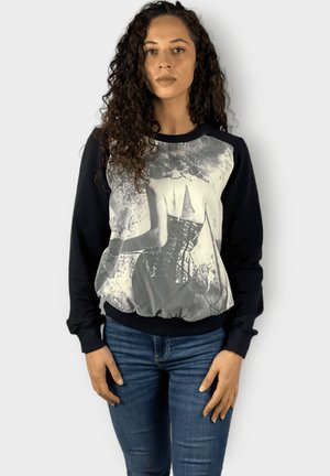 Mujer con cabello rizado que lleva puesta una sudadera negra con una impresión artística en escala de grises de una mujer en corsé, combinada con unos jeans azules.