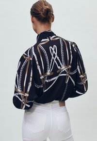 Czarna bluzka z abstrakcyjnym białym i złotym nadrukiem w kształcie szelki, luźny krój, długie rękawy, marszczone ramiona i gładka tekstura materiału.