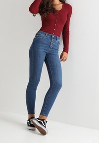 Korkeavyötäröiset siniset skinny-farkut, joissa on viiden napin sulku, valmistettu denimkankaasta, yhdistetty burgundinpunaisiin pitkähihaisiin paitaan ja mustiin lenkkareihin.