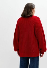 Röd oversize stickad tröja med texturerad tyg, ribbade muddar och nederkant. Kombinerad med blå denimbyxor. Bakvy.