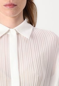 Camicia bianca, leggera e trasparente, con pieghe verticali, colletto classico e patta con bottoni, che enfatizza una texture leggera e ariosa.