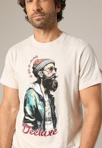 T-shirt en coton beige avec un graphique coloré d'un homme barbu, portant des lunettes de soleil et un bonnet en tricot, avec le texte "Deeluxe."