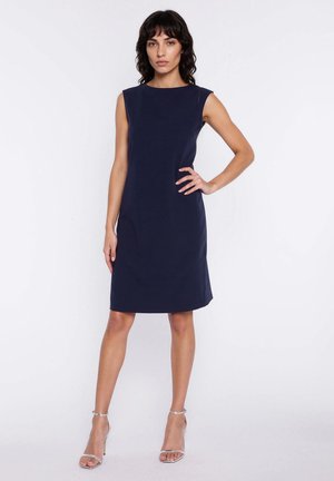 Kobieta stojąca z ręką na biodrze, ubrana w granatową, bezrękawnikową sukienkę do kolan oraz srebrne sandały na wysokim obcasie, na jednolitym białym tle.
