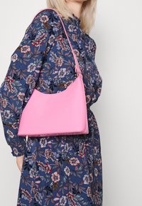 Sac à main rose brillant avec une ligne supérieure incurvée et une courte bandoulière, présenté sur une robe à imprimé floral. Design lisse et minimaliste.