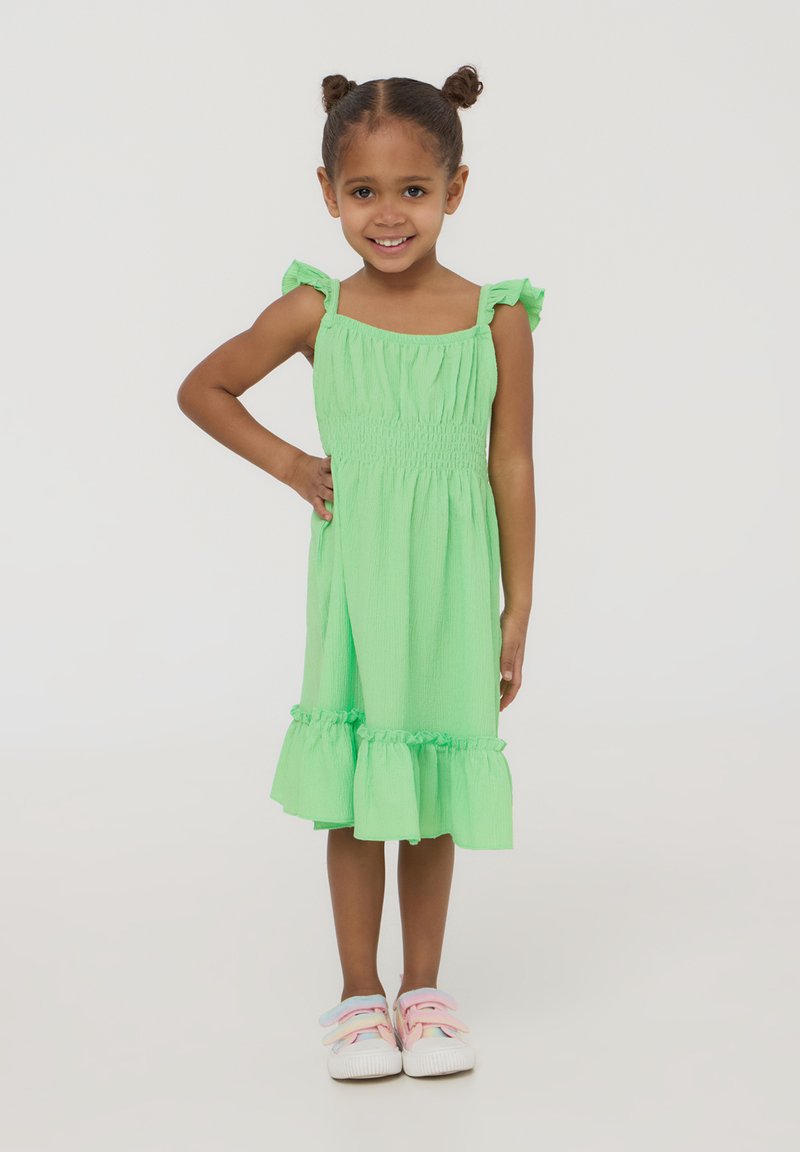 Abito di cotone verde con corpino arricciato, spalline a volant e orlo svasato. Abbinato a scarpe bianche con cinturini color pastello.