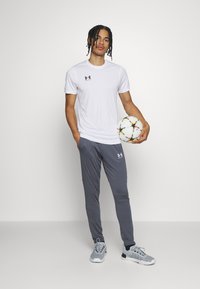 Balti sportiniai marškinėliai su juodu logotipu, derinami su pilkomis sportinėmis kelnėmis. Modelis laiko spalvingą futbolo kamuolį ir vilki pilkus sportinius batelius.