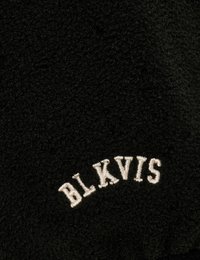 Svart fleece-tyg med en böjd, vit broderad text "BLKVIS" i nedre högra hörnet. Mjuk textur med en fluffig utseende.