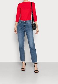 Rotes langärmliges Oberteil kombiniert mit hoch taillierten blauen Jeans. Schwarze Riemchensandalen und eine kleine schwarze Umhängetasche runden den Look ab.